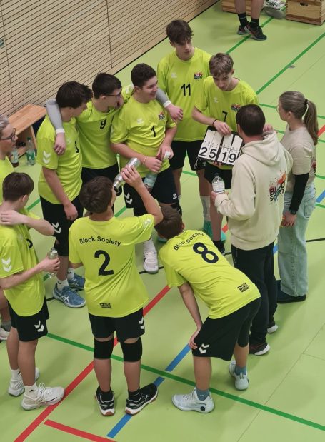 Junge Volleyballmannschaft in grünen Trikots bespricht Taktiken in einer Sporthalle.